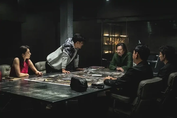 The cast of Tokyo Swindlers engaged in an intense discussion around a large table.