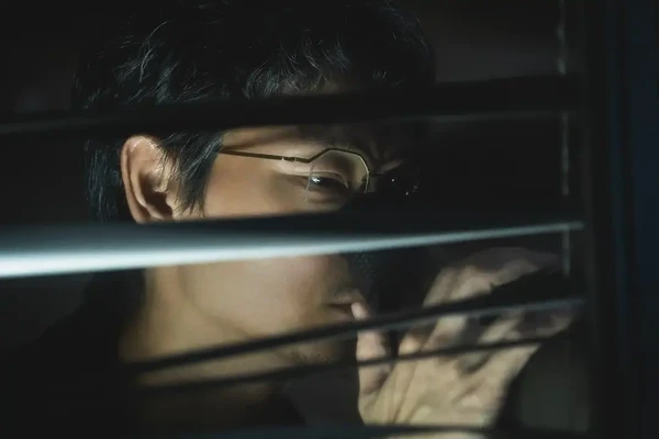 A character in Tokyo Swindlers secretly peers through blinds, suggesting a sneaky act.